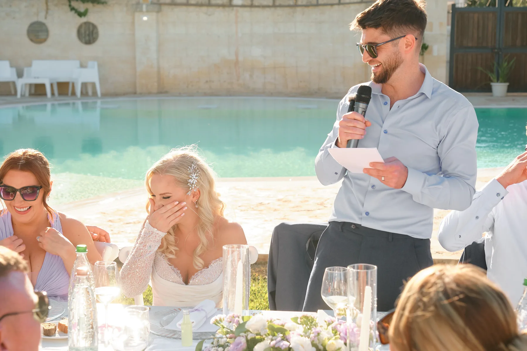 Lo sposo legge il suo discorso al microfono mentre la sposa ride commossa, a bordo piscina